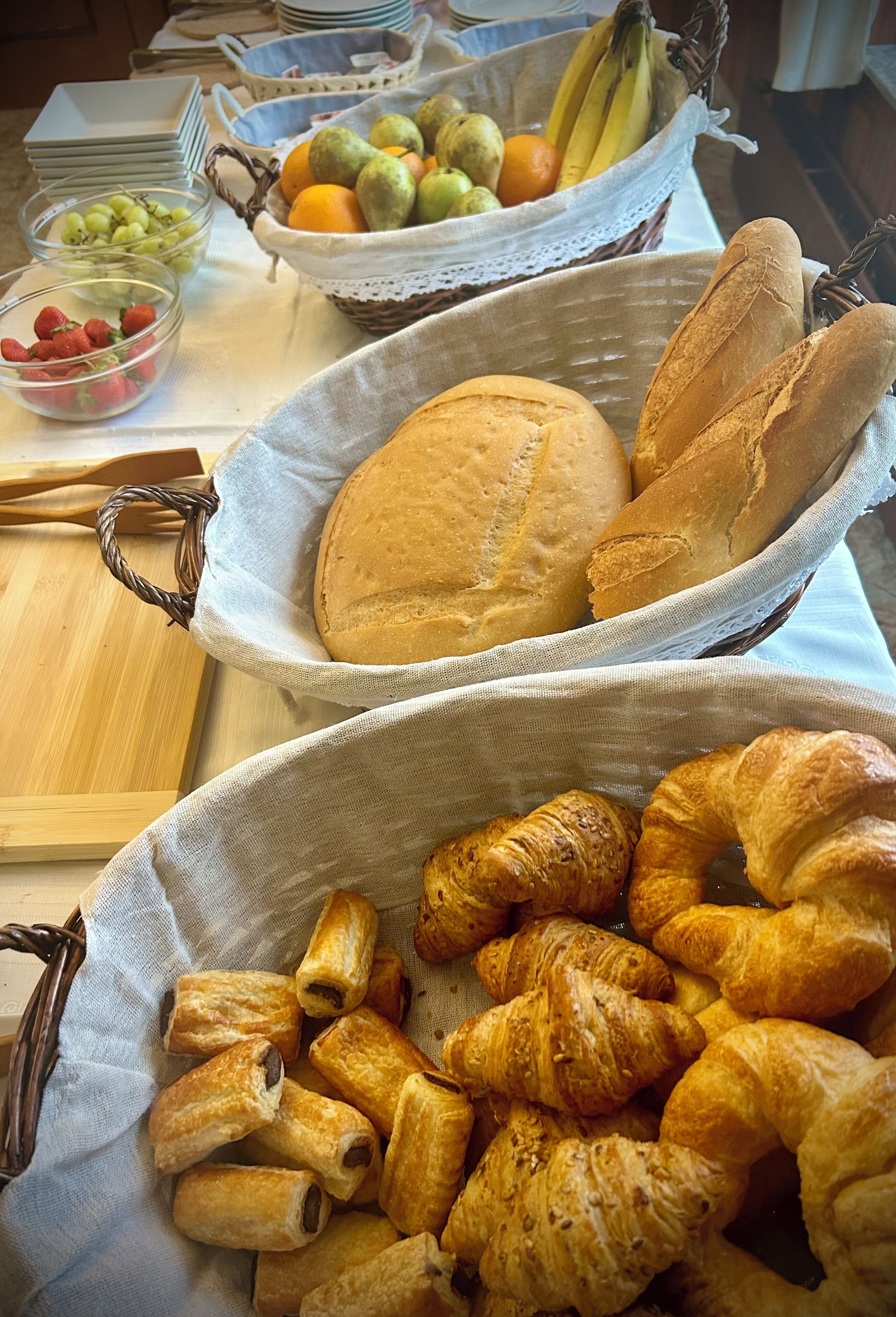Une table sur laquelle on trouve des paniers de pain et de viennoiseries, des saladiers remplis de fruits et une pile d’assiettes.