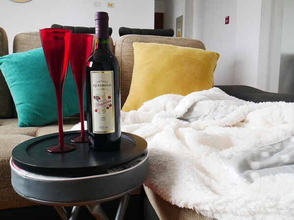 A bottle of wine on a coaster next to a red wine glass on a small table in a living room with a sofa and colorful cushions.