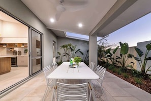 A modern outdoor dining area with a white table and chairs, a glass door leading to a kitchen, and tropical plants. - The Indiana A Space to Relax (Dunsborough)