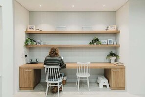A modern workspace with a wooden desk, two white chairs, and shelves with plants and pictures.