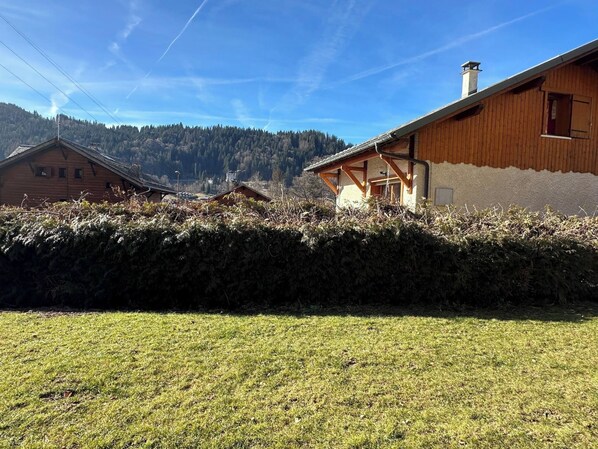 A wooden house with a stone wall in front, a grassy yard, and a forested mountain in the background.