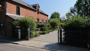 A brick building with a gated entrance, a gravel driveway, and a metal fence. - Gîte la Roseraie (Beuvron-en-Auge)