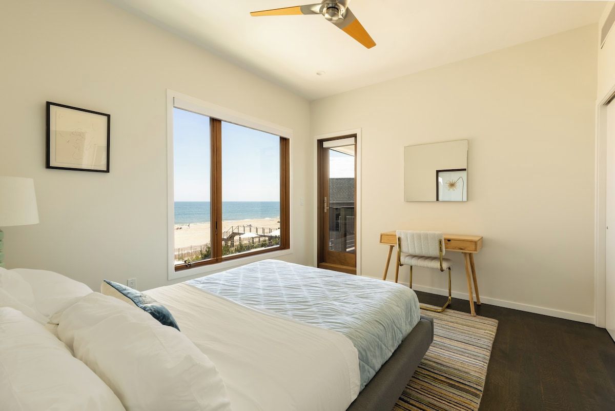 A bedroom with a bed, a standing desk, a chair, a ceiling fan, a large window with a beach view, and a framed picture on the wall.
