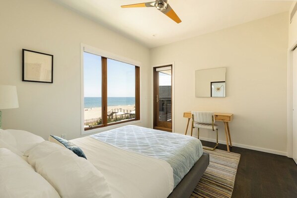 A bedroom with a bed, a standing desk, a chair, a ceiling fan, a large window with a beach view, and a framed picture on the wall.
