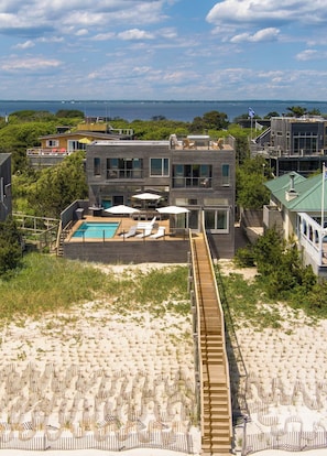 A modern two-story house with a pool, surrounded by a wooden deck and beach chairs.