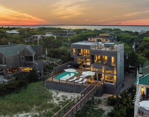 A modern multi-story house with a swimming pool, surrounded by greenery and a view of the water.