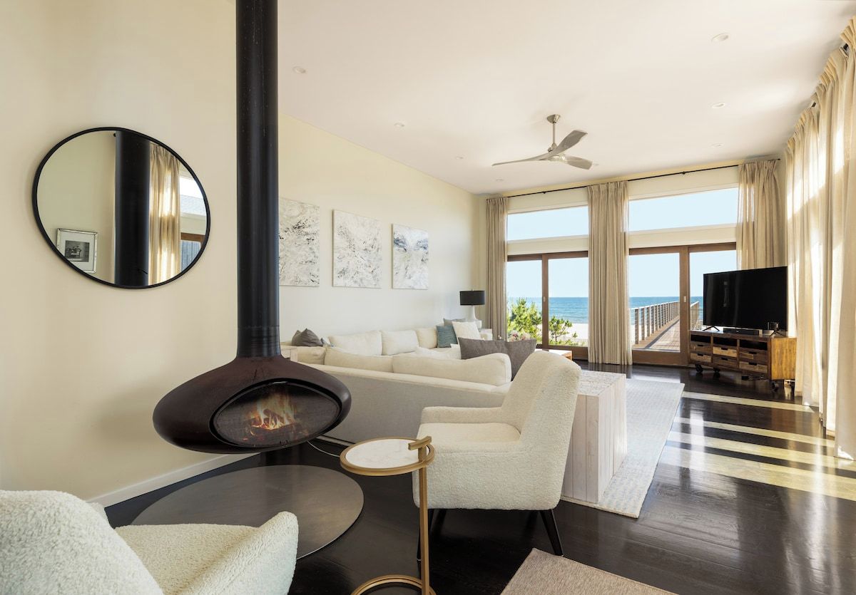 A modern living room with a fireplace, a sofa set, a flat-screen TV, and a view of the ocean.