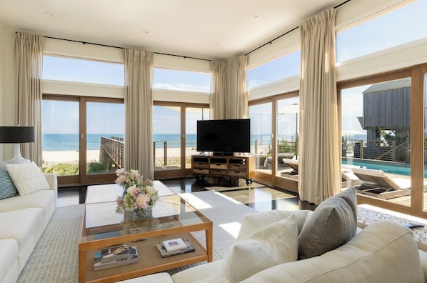 A modern living room with a large TV, comfortable seating, a coffee table, and a view of the ocean.