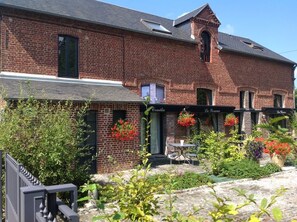 A brick building with a garden patio area, outdoor seating, and hanging flower baskets. - Gîte le Camélia (Beuvron-en-Auge)