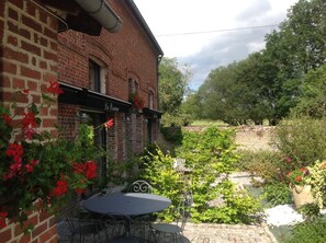 A brick building with a patio area featuring a table and chairs, surrounded by lush greenery and flowering plants. - Gîte le Camélia (Beuvron-en-Auge)