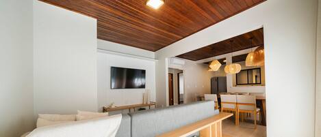 A modern living room with a wooden ceiling, a flat-screen TV mounted on the wall, a grey sofa, and a wooden coffee table with books.
