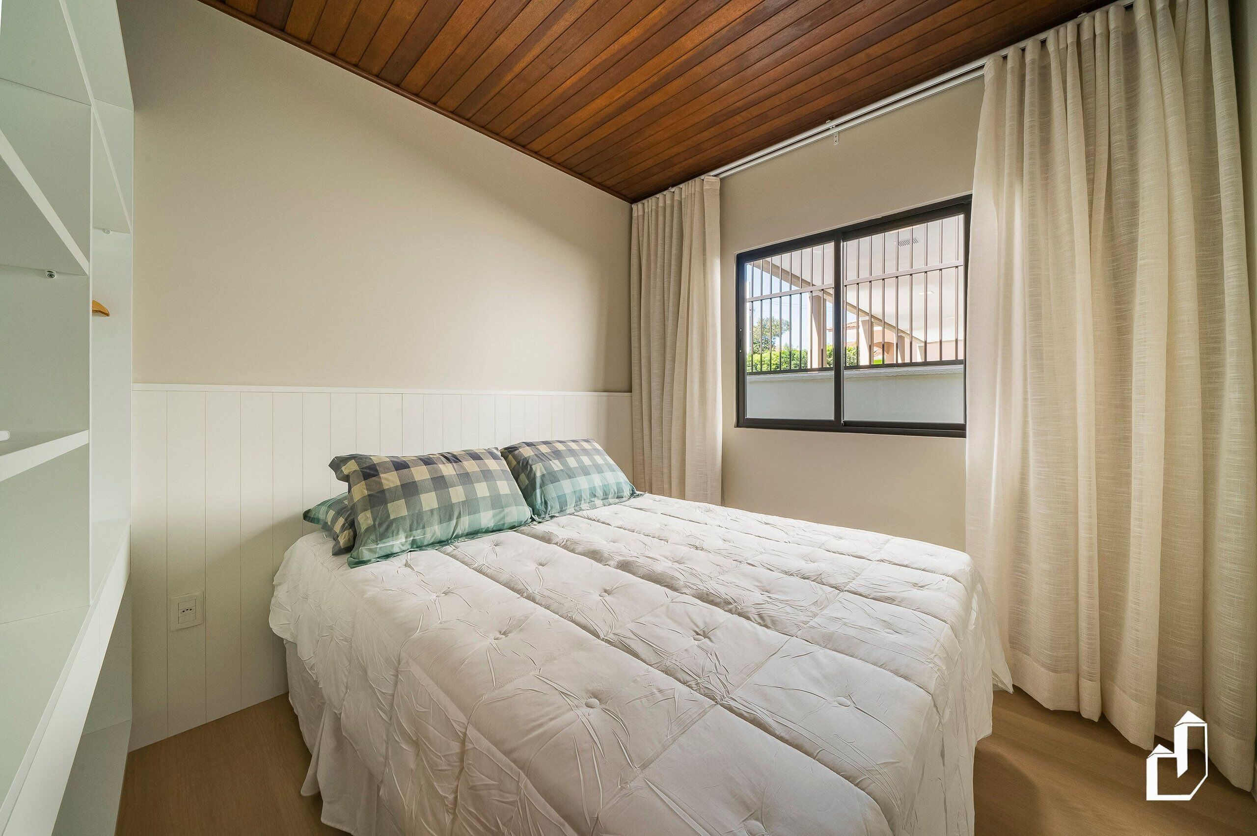 Un dormitorio con cama, ventana con cortinas, techo de madera y pared blanca.