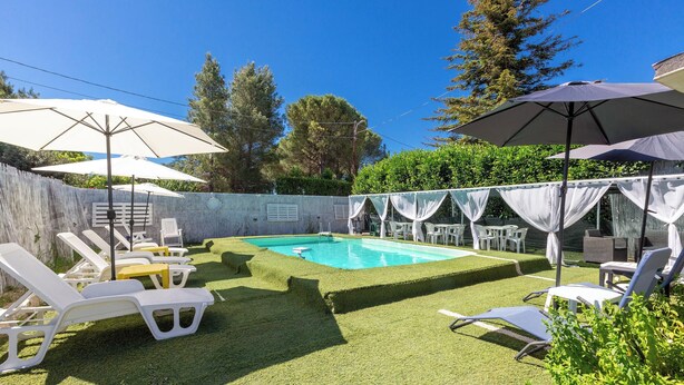 Ein Garten mit Swimmingpool, umgeben von weißen Liegestühlen und Sonnenschirmen, sowie ein überdachter Sitzbereich.