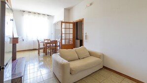 Ein Wohnzimmer mit einem beigen Sofa, ein Essbereich mit einem Holztisch und Stühlen sowie eine durch eine offene Tür einsehbare Küche.