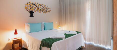 Una cama bien hecha con ropa de cama blanca y verde, una mesita de noche con una lámpara, un aire acondicionado instalado en la pared y una ventana con cortinas traslúcidas.