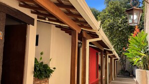 Un pasillo exterior cubierto con vigas de madera, plantas en macetas y un muro de ladrillo.