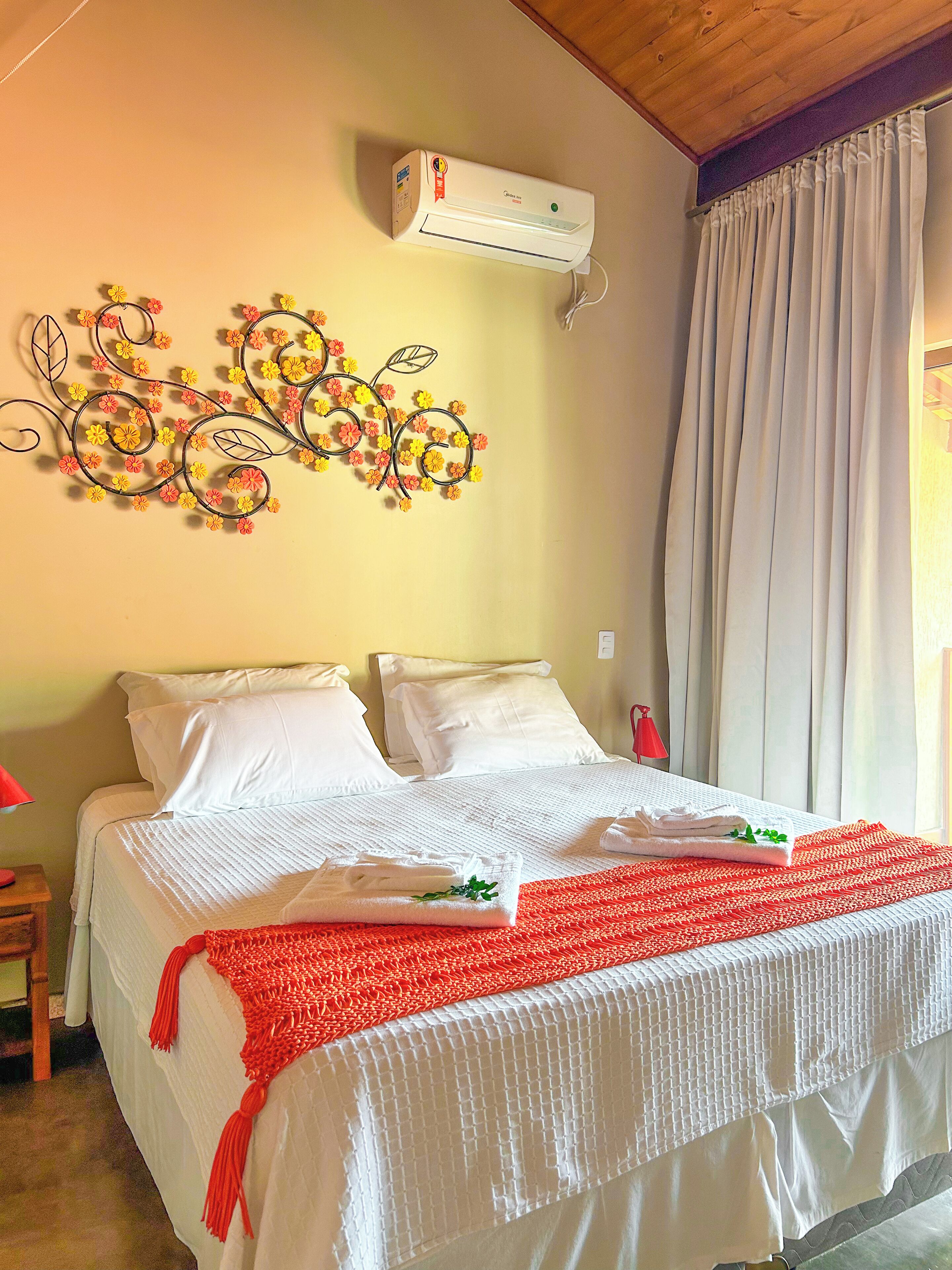 A neatly made bed with white linens and a red blanket, a wall-mounted air conditioner, and a decorative floral wall art piece.