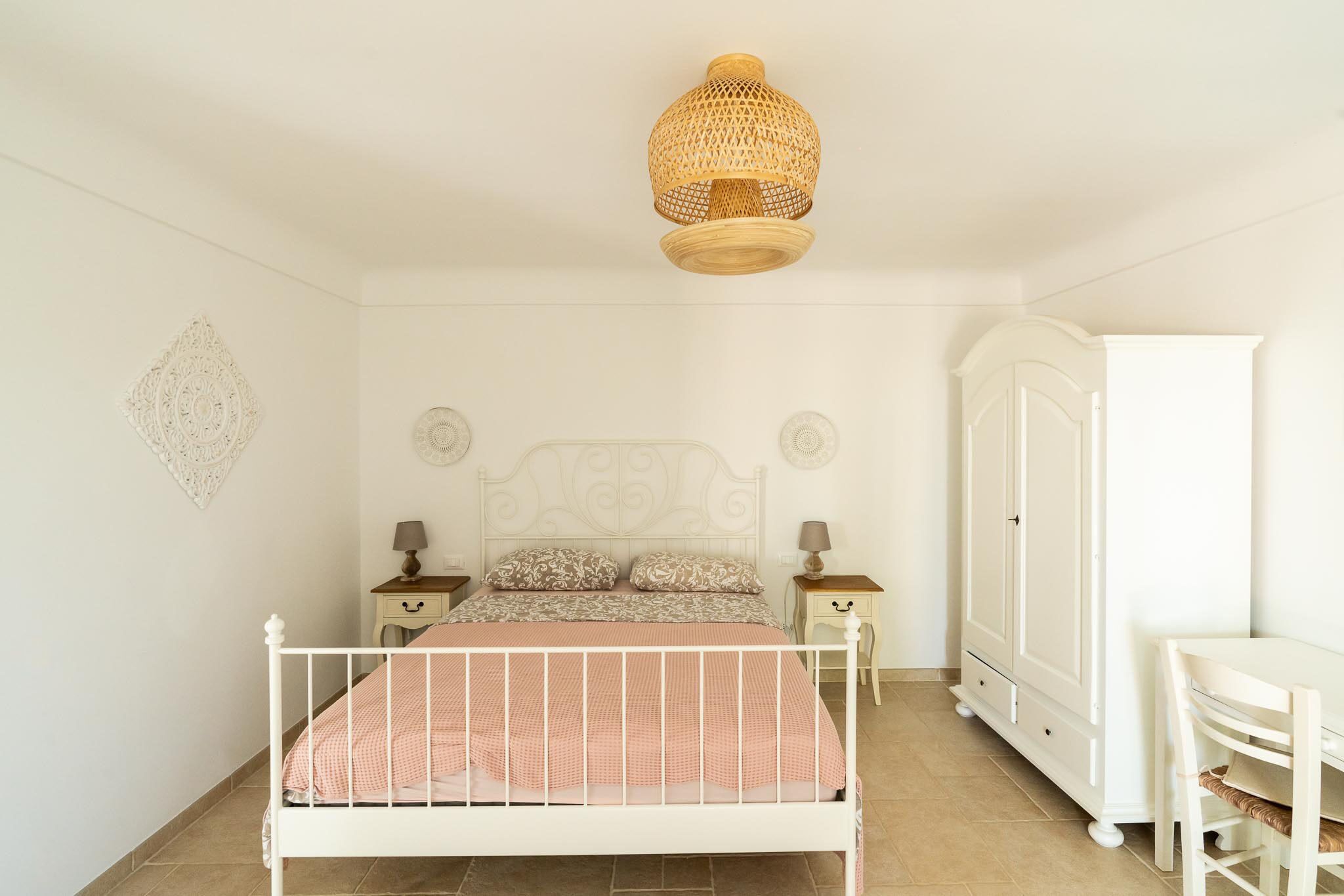 Ein Schlafzimmer mit weißem Metallbettgestell, einem weißen Kleiderschrank, einem Nachttisch mit Lampe sowie einem Wandbild.