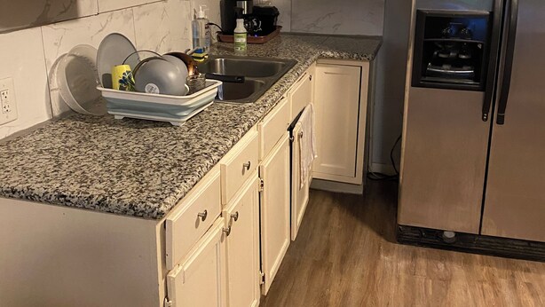 Una cocina con armarios blancos, encimera de granito, refrigerador de acero inoxidable y suelo de madera.