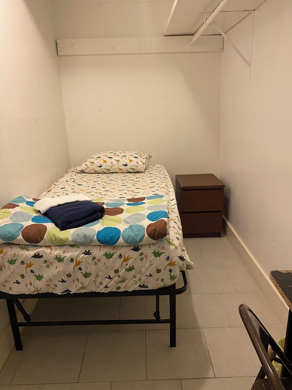 A small, single-bed room with a patterned bedspread, a pillow, and a bedside table with a drawer.