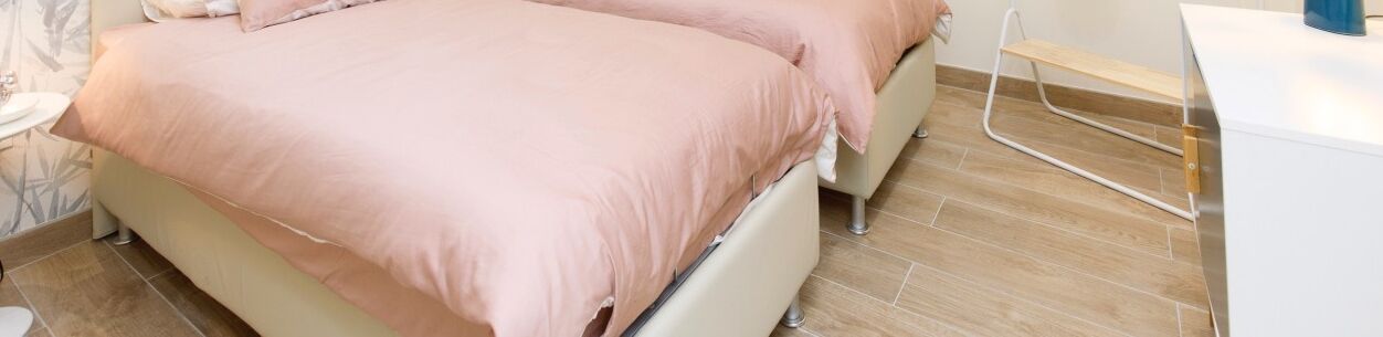 Een bed met een roze sprei in een kamer met een betegelde vloer en een witte nachtkastje.