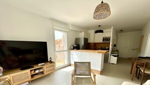 Uma sala de estar moderna com televisão de tela plana, móveis de madeira, área de cozinha com armários brancos e uma grande janela que oferece iluminação natural.