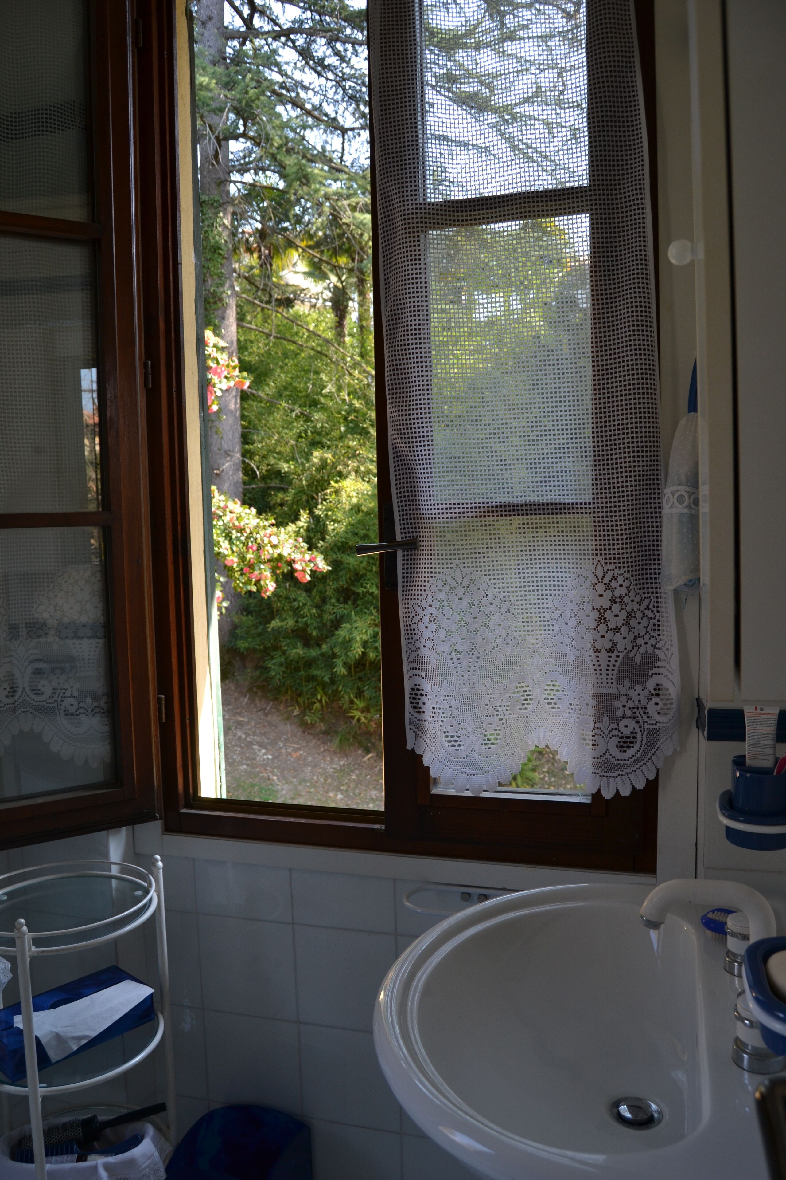 Een badkamer met een wastafel, een raam met kantgordijnen en uitzicht op bomen.