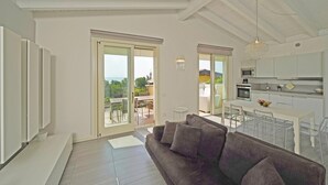 A modern living room with a grey sofa, a dining area with a white table and chairs, and a kitchen with white cabinets and a hanging light fixture. - Montecolo Resort E7 by Wonderful Italy (Manerba del Garda)