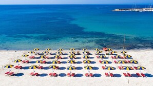 A beach with rows of sunbeds and umbrellas, a clear blue sea, and a pier in the distance.