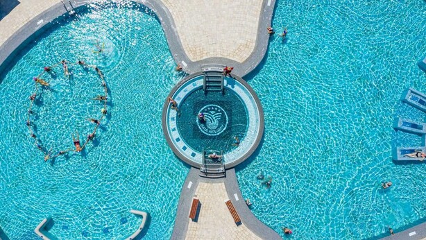 An aerial view of a swimming pool with a central circular area marked by a logo, surrounded by a tiled walkway and lounge chairs.