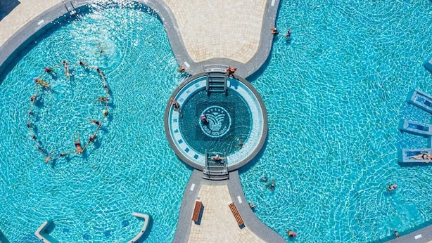 Vista aérea de una piscina con un área circular central marcada por un logotipo, rodeada por un paseo de baldosas y sillas de descanso.