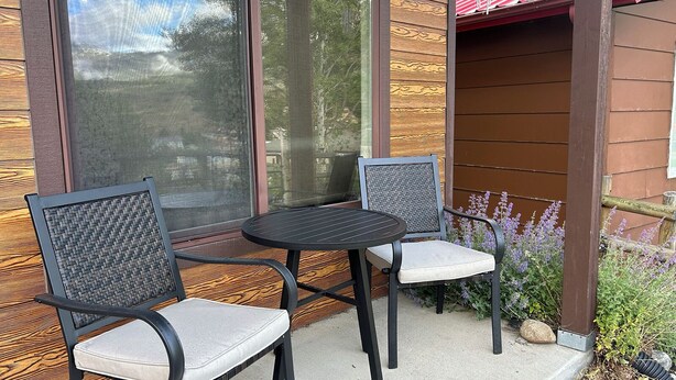 A patio with two chairs and a table, a wooden exterior wall, and a window with a view of trees.