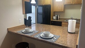 A kitchen with granite countertops, wooden cabinets, a refrigerator, a coffee maker, and a paper towel dispenser.