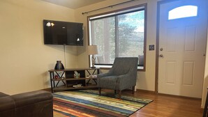 A living room with a wooden floor, a gray armchair, a TV mounted on the wall, and a window with a view of trees.