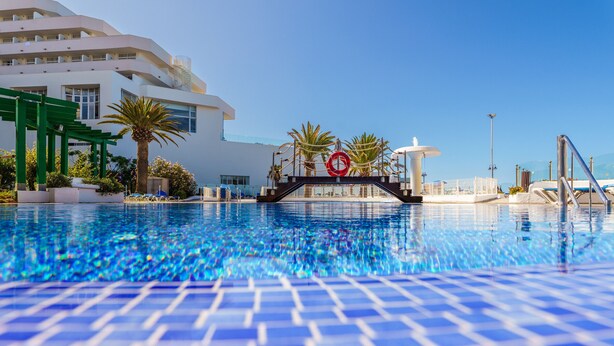 A swimming pool with a clear blue tile pattern, surrounded by palm trees and a modern building with balconies. - Exclusive Sea Views Villamar - Big Terrace & Pool (Costa Adeje)