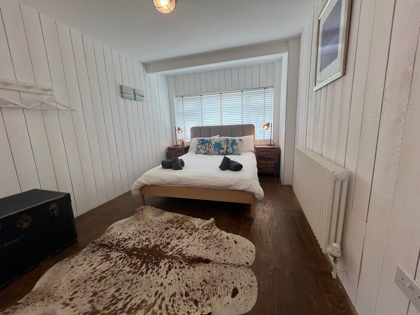 A bedroom with a bed, bedside tables, a radiator, and a window with blinds.