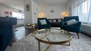 Ein modernes Wohnzimmer mit türkisfarbenen Ohrensesseln, einem Glastisch und einer Couch mit gemusterten Kissen.