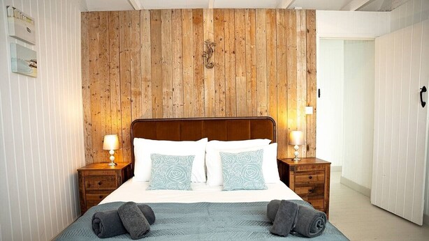 Ein Schlafzimmer mit einem hölzernen Kopfteil, zwei Nachttischen mit Lampen, einem Bett mit weiß-blauen Kissen und einer blauen, strukturierten Tagesdecke.