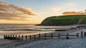 A coastal landscape with a wooden walkway leading to a beach, a cliff in the background, and a sunset sky.