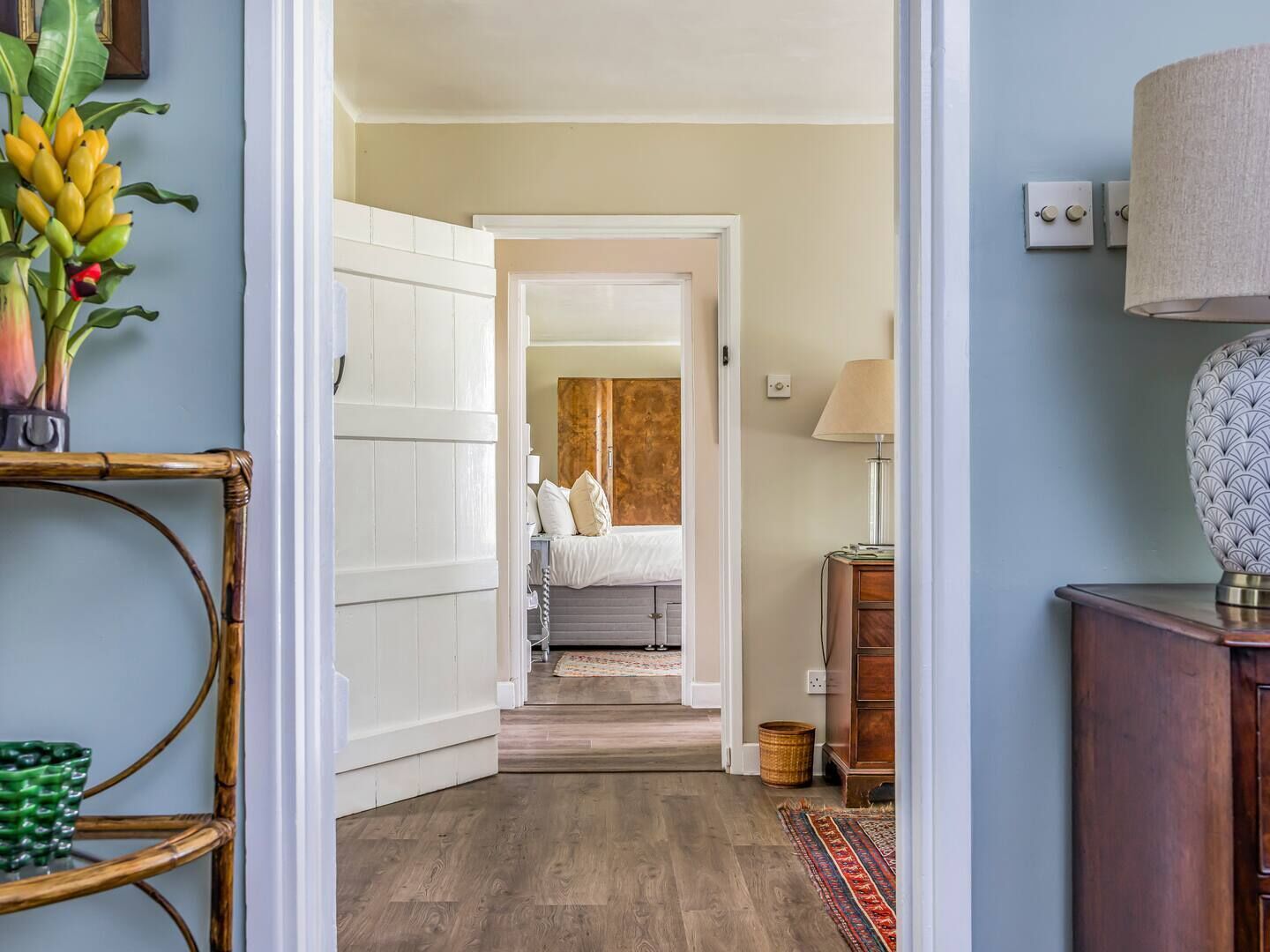 A hallway leading to a bedroom with a bed, bedside table, lamp, and a decorative plant.