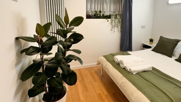 A modern bedroom with a single bed, a potted plant, a window with curtains, a radiator, and a bedside table with a lamp. - Cosy Apartment in Canary Wharf  (London)
