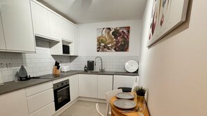 A compact kitchen with white cabinets, a built-in oven, a sink, and a dining area with a wooden table and chairs. - Cosy Apartment in Canary Wharf  (London)