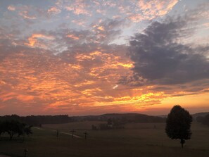Un coucher de soleil sur un paysage rural, avec des champs, quelques arbres et des collines à l’horizon.
