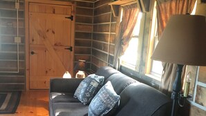 A cozy cabin interior with a wooden floor, a dark sofa with patterned cushions, a lamp, and a basket with a knitted item.