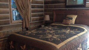 A log cabin bedroom with a bed, a window with curtains, a nightstand, a lamp, and a framed picture on the wall.