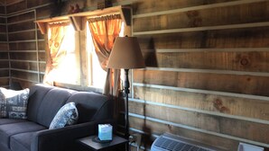 A cozy cabin interior with a wooden ceiling, a sofa with cushions, a table with a lamp, and an air conditioning unit.