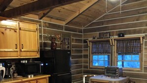 A cabin interior with wooden walls, a kitchen area including a refrigerator, a dining table, and a window with plaid curtains.