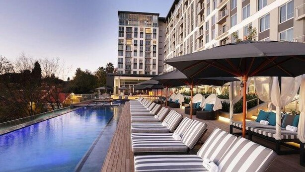 A hotel pool area with sun loungers and umbrellas, a clear pool, and a high-rise building in the background. - Sandtoncity-apartment-sleeps4-pool-parking (Johannesburg)