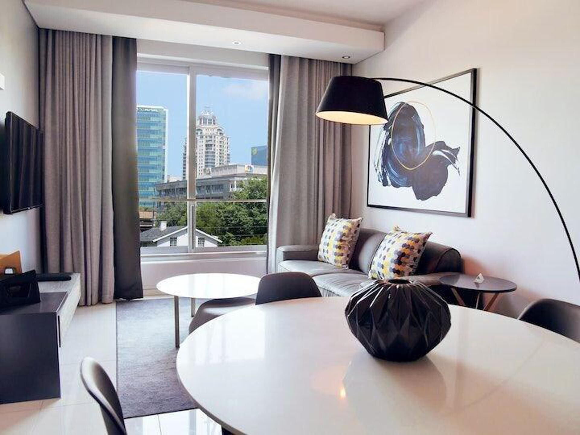 Una habitación de hotel moderna con comedor, un sofá con cojines estampados, un ventanal con vistas a la ciudad y un cuadro en la pared.