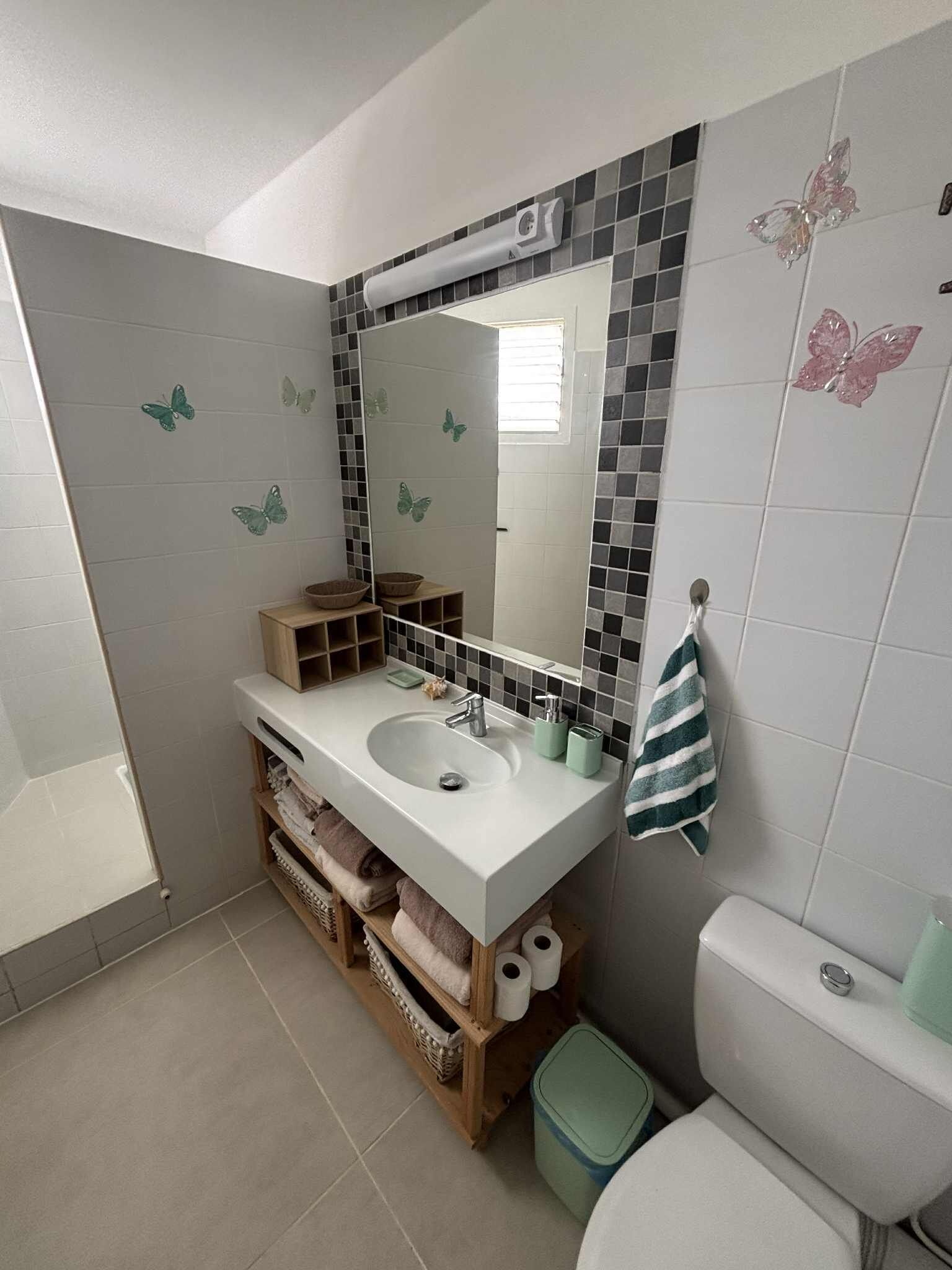 Een badkamer met een witte wastafel, spiegel en toilet. Op de muur zitten vlinderstickers, er is een handdoekhouder en een prullenbak.